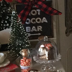 May include: A miniature Christmas scene with a sign that reads "Hot Cocoa Bar" with a red and black plaid bow. There is a small Christmas tree, a glass jar filled with peppermint candies, and a cake under a glass dome.
