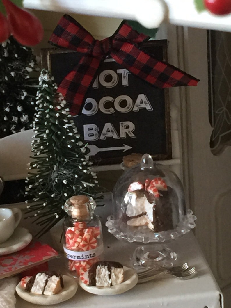 May include: A miniature Christmas scene with a sign that reads "Hot Cocoa Bar" with a red and black plaid bow. There is a small Christmas tree, a glass jar filled with peppermint candies, and a cake under a glass dome.