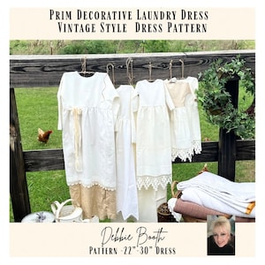 以下が含まれることがあります： 木製の柵に掛けられた、いくつかの白いヴィンテージスタイルのドレス。 ドレスにはレースと生地のディテールがあります。 画像のテキストは「PRIM DECORATIVE LAUNDRY DRESS VINTAGE STYLE DRESS PATTERN」と「Debbie Booth PATTERN -22"-30" DRESS」です。