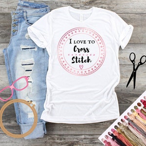 May include: White t-shirt with a pink circle design that says "I Love To Cross Stitch" with a pink heart. The shirt is laying on a wooden surface with a pair of scissors, a wooden embroidery hoop, and a variety of embroidery floss.