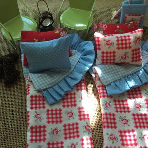 May include: Two red and white gingham sleeping bags with blue and white ruffled edges and blue polka dot pillows. The sleeping bags are on a burlap surface with two green metal chairs and a lantern.