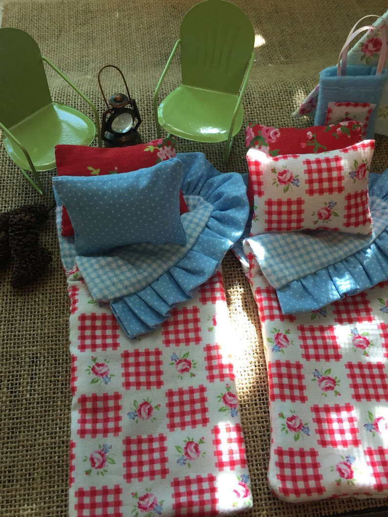 May include: Two red and white gingham sleeping bags with blue and white ruffled edges and blue polka dot pillows. The sleeping bags are on a burlap surface with two green metal chairs and a lantern.