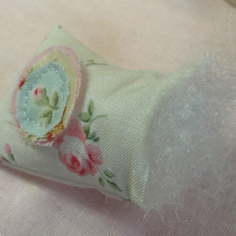 Op de afbeelding: Een klein, wit kussen met een bloemenpatroon en een lichtblauwe en roze bloemenapplicatie. Het kussen is gevuld met witte vulling en heeft een rauwe rand.