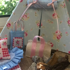 May include: A miniature blue and white gingham tent with a pink and white gingham sleeping bag, two blue and white gingham bags, and a pink and white gingham tote bag. There are also small rocks and a piece of driftwood in the foreground.