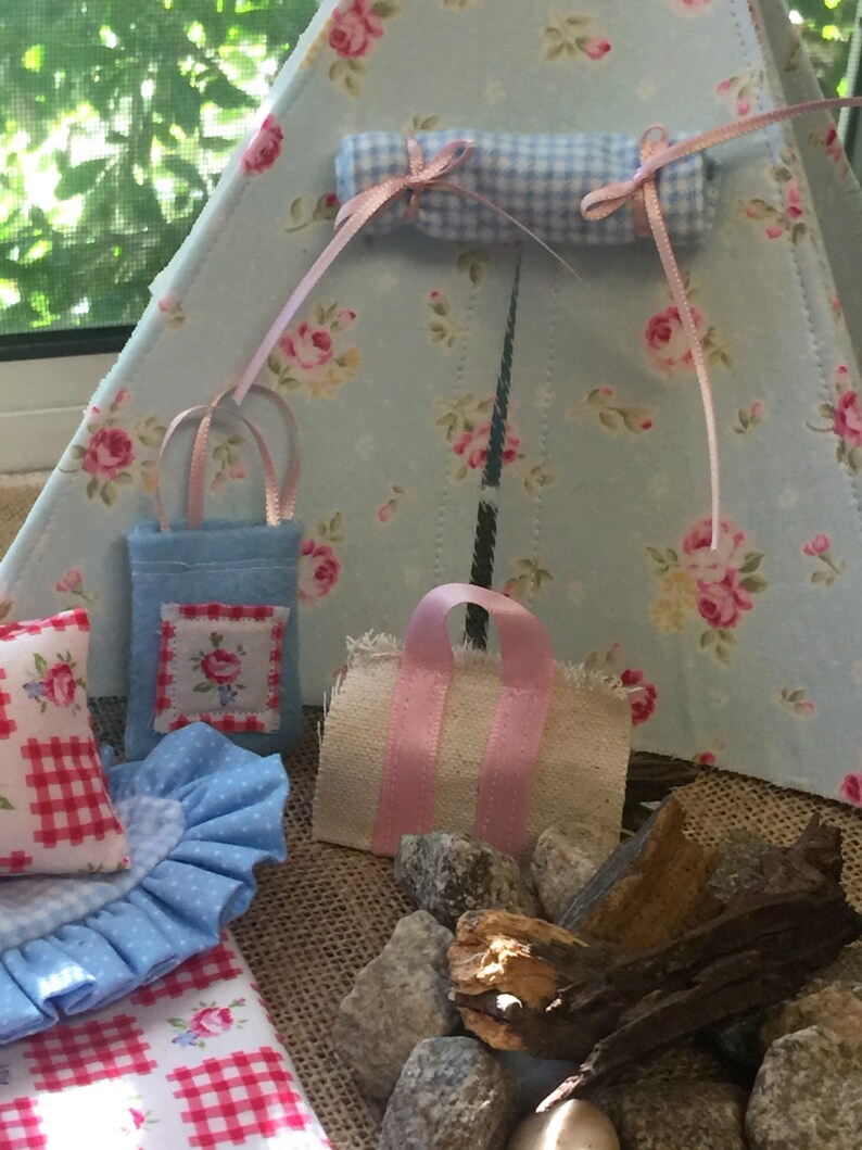 May include: A miniature blue and white gingham tent with a pink and white gingham sleeping bag, two blue and white gingham bags, and a pink and white gingham tote bag. There are also small rocks and a piece of driftwood in the foreground.