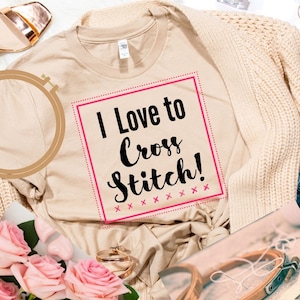 May include: A beige t-shirt with the text "I Love to Cross Stitch!" in black lettering inside a pink square with pink x's around the border. The shirt is lying on a white surface with a wooden embroidery hoop, a pair of gold sandals, and a beige cardigan.