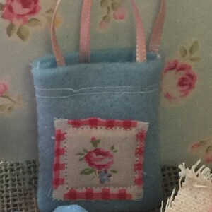 May include: A small blue felt bag with a pink ribbon handle and a white and red embroidered floral patch. The bag is perfect for small gifts or trinkets.