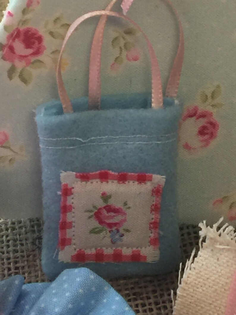 May include: A small blue felt bag with a pink ribbon handle and a white and red embroidered floral patch. The bag is perfect for small gifts or trinkets.