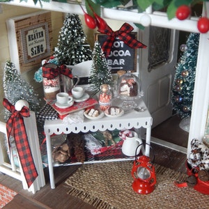 May include: A miniature dollhouse scene with a white picket fence, a red and black checkered bow, a small table with a red lantern, a sign that says "Hot Cocoa Bar", and a basket of pine cones.
