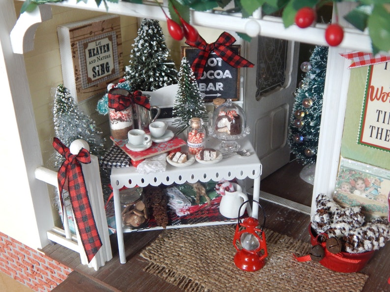 May include: A miniature dollhouse scene with a white picket fence, a red and black checkered bow, a small table with a red lantern, a sign that says "Hot Cocoa Bar", and a basket of pine cones.