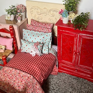 May include: A red and white checkered doll bed with a floral patterned comforter and pillows. The bed is set up in a dollhouse room with a red cabinet and a white headboard.