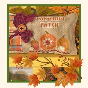 Pode incluir: Uma almofada bege com um design de ponto cruz com abóboras e o texto "Pumpkin Patch". A almofada é decorada com um laço de tecido bordô e um botão. A almofada está sentada em um banco amarelo com folhas de outono e bagas.