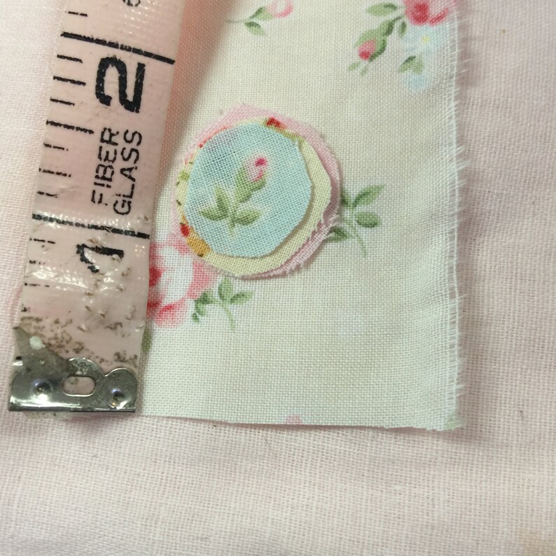 Op de afbeelding: Een close-up van een stuk stof met een bloemenpatroon en een gelaagde stofbloem applicatie in tinten roze, geel en blauw.