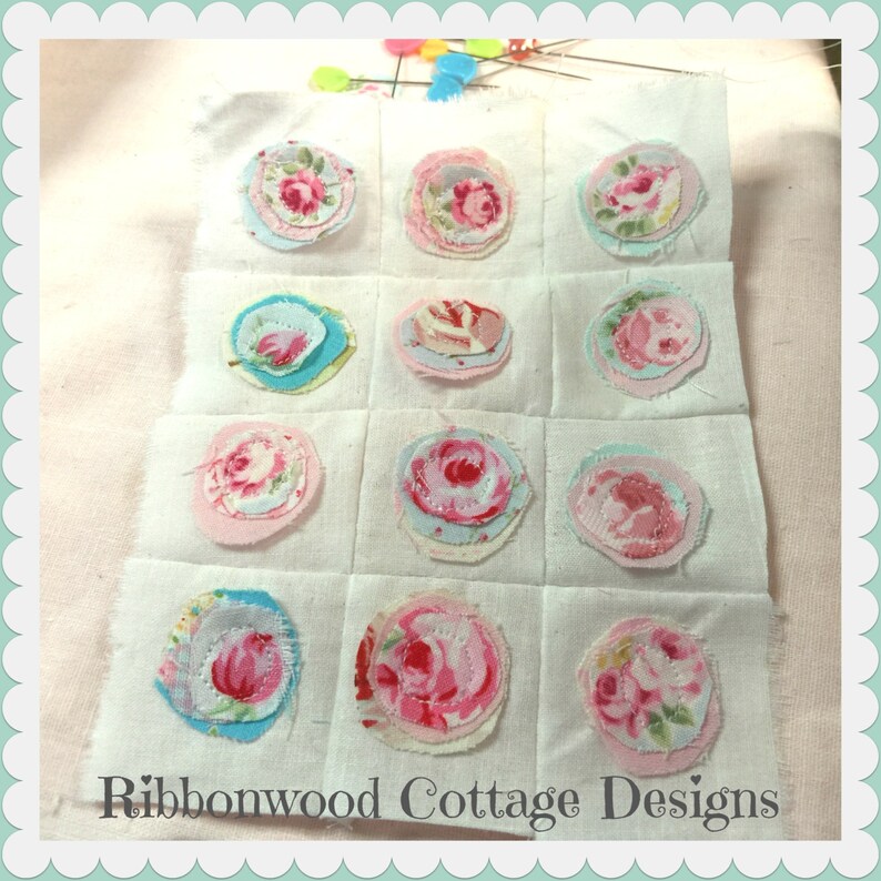 Op de afbeelding: Een wit vierkant van stof met 12 ronde stoffen applicaties in tinten roze, blauw en wit. Elke applicatie is gemaakt van meerdere lagen stof en heeft een bloemenmotief. Het vierkant van stof is gelabeld "Ribbonwood Cottage Designs".