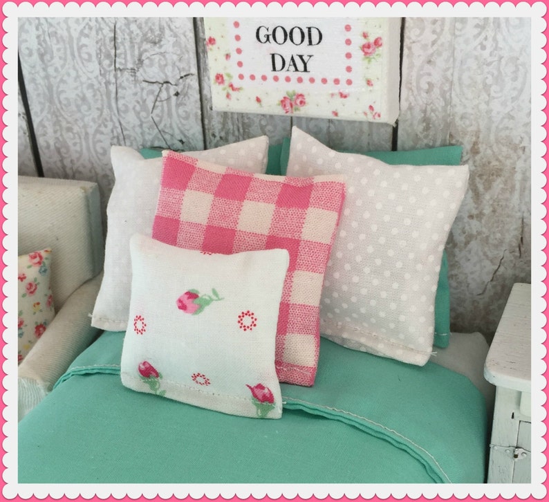 May include: A bed with a teal green duvet cover and three pillows. The pillows are white with pink and white gingham, white with white polka dots, and white with pink floral designs. The sign above the bed reads "Good Day".