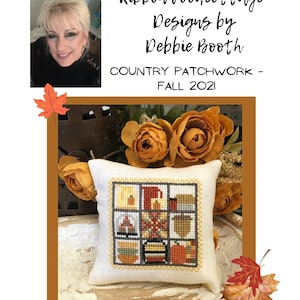 May include: A small white pillow with a cross-stitch design featuring a patchwork of autumnal scenes. The pillow is surrounded by yellow flowers and fall leaves.