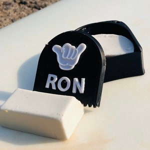 May include: A black surfboard wax container with a white hand symbol and the word 'RON' in white letters. The container is open and a white surfboard wax bar is next to it.