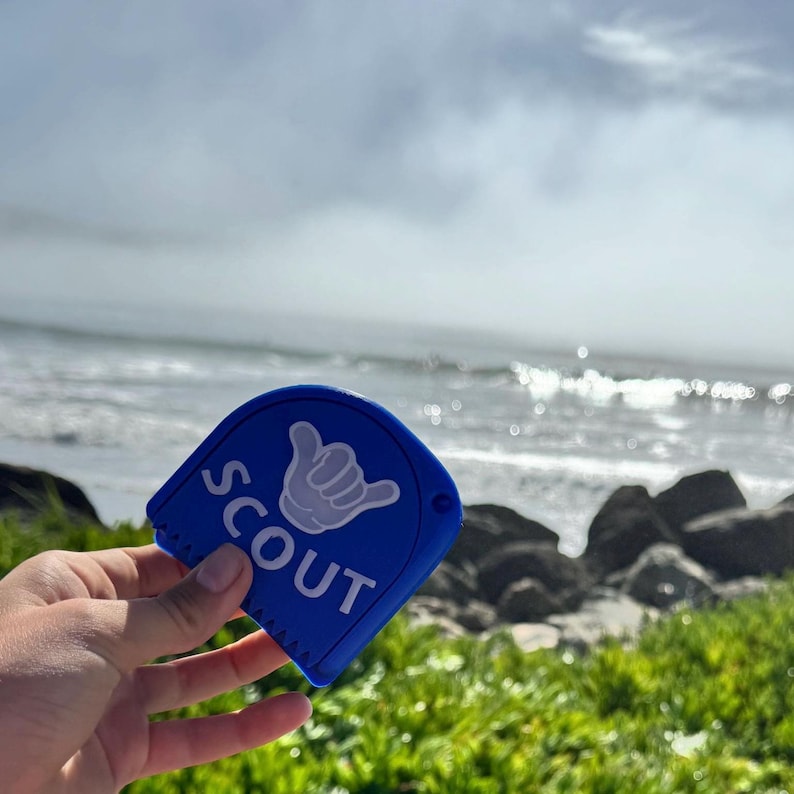 May include: Blue plastic surfboard wax comb with a white "SCOUT" logo and a white hand gesture graphic. The comb is being held in front of a beach scene with waves and rocks.
