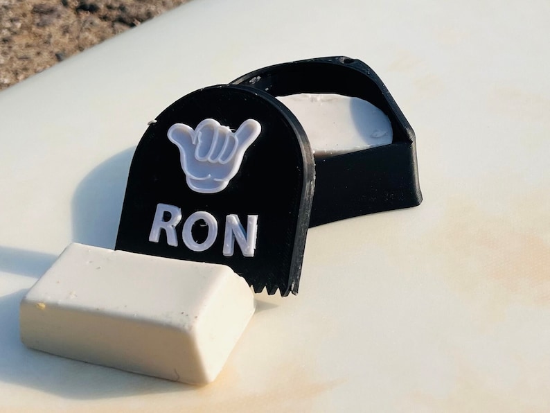 May include: A black surfboard wax container with a white hand symbol and the word 'RON' in white letters. The container is open and a white surfboard wax bar is next to it.