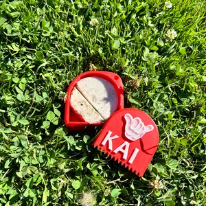 May include: Red plastic container with a white surfboard wax inside. The container has a white hand symbol and the word "KAI" on the front.