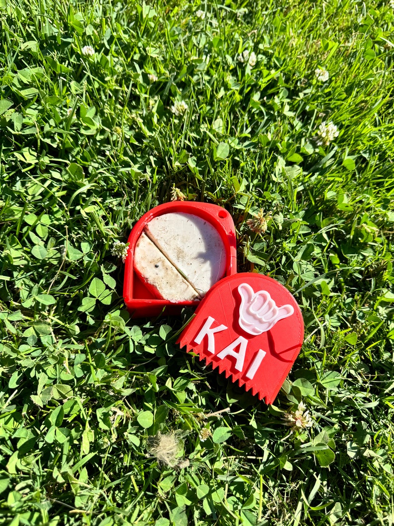 May include: Red plastic container with a white surfboard wax inside. The container has a white hand symbol and the word "KAI" on the front.