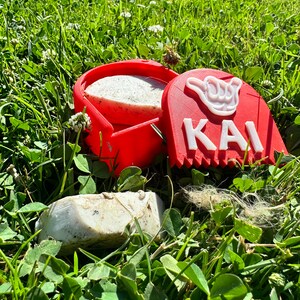 May include: A red plastic container with a white soap bar inside. The container has a white hand symbol and the word "KAI" on the side. The container is sitting on green grass.