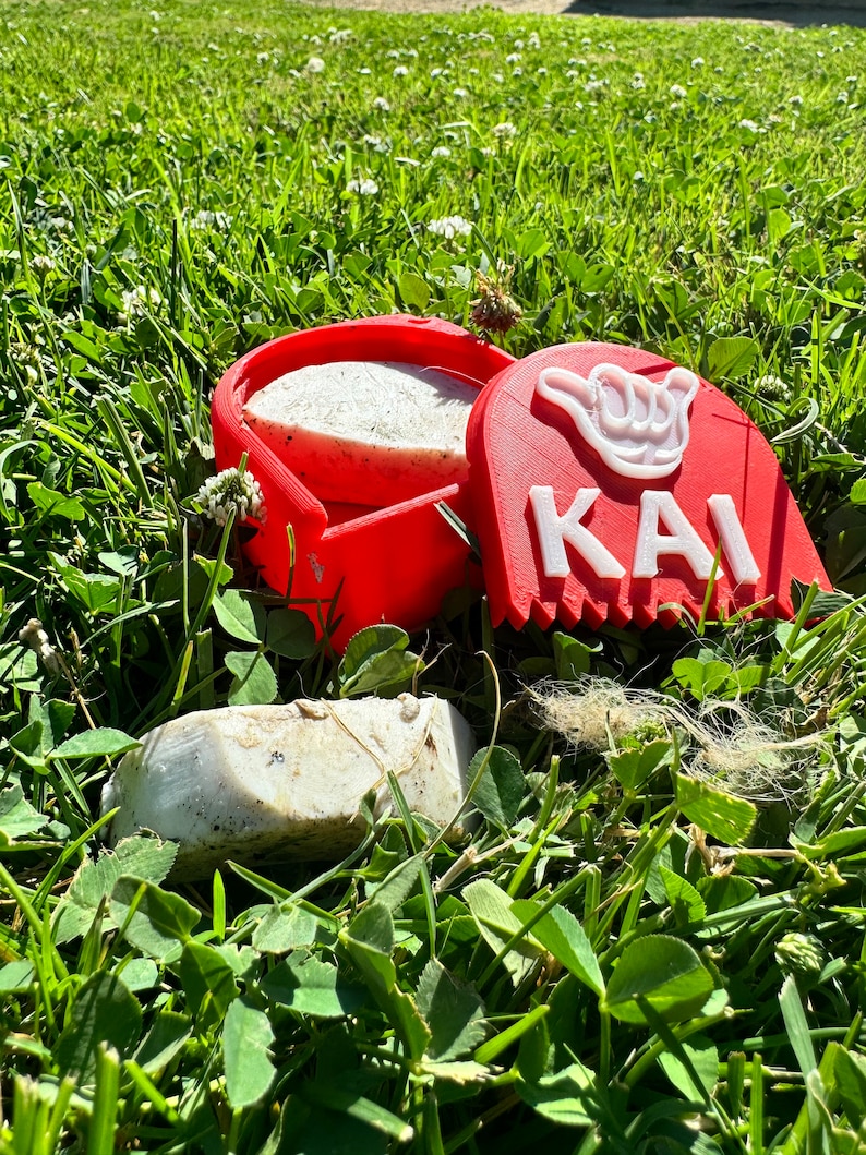 May include: A red plastic container with a white soap bar inside. The container has a white hand symbol and the word "KAI" on the side. The container is sitting on green grass.