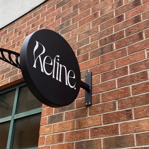 Puede incluir: Un letrero redondo negro con texto blanco que dice "Refine" montado en una pared de ladrillo.