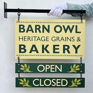 May include: A sign for "Barn Owl Heritage Grains & Bakery" with "Open" and "Closed" options. The sign is cream-colored with dark green text and a black metal bracket. The sign is held by a person wearing a white glove.