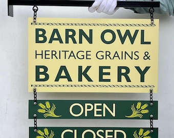 Peut inclure: Enseigne de boulangerie "Barn Owl Heritage Grains & Bakery" avec texte vert foncé sur fond crème. Les panneaux "Ouvert" et "Fermé" sont suspendus. Le tout est maintenu par une main gantée et un support en métal noir.