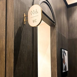 May include: A round, white sign with a gold trim and a brass bracket attached to a dark wood wall. The sign has a symbol for men's and women's restrooms and a wheelchair symbol. The sign also says "WC".
