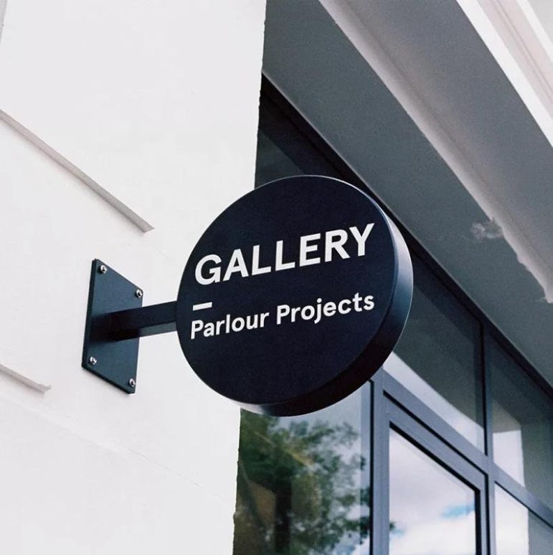 May include: A black circular sign with white text that reads "GALLERY - Parlour Projects" hanging on a white building.