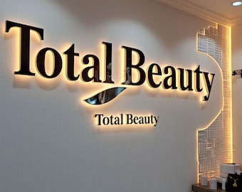 May include: A sign for "Total Beauty" on a wall, featuring black lettering with a warm, illuminated backlight. The sign is mounted on a light-coloured wall, with a smaller "Total Beauty" sign below.