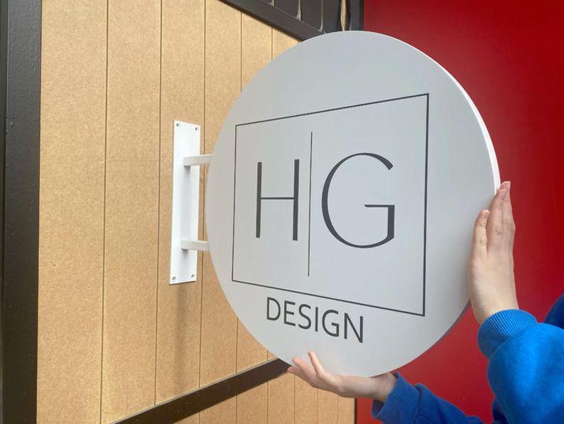 May include: White round sign with black text that reads "HG DESIGN". The sign is mounted to a wall with a white bracket.