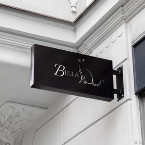 May include: A black sign with white lettering and a silhouette of a cat. The sign reads "Bella".