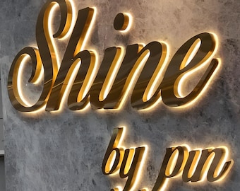Puede incluir: Letrero dorado e iluminado que dice "Shine by pun" en una fuente cursiva. El letrero está montado en una pared gris texturizada. Las letras tienen una luz cálida y brillante que las delinea.