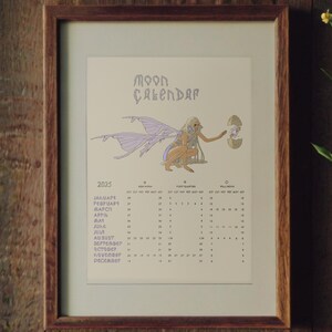 May include: Framed 2025 Moon Calendar print. The artwork features a whimsical illustration of a winged creature holding a lantern. The calendar is in a wooden frame, set against a wood-paneled background. A sprig of greenery with yellow flowers is to the right.