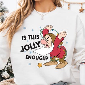 May include: White sweatshirt with the phrase "IS THIS JOLLY ENOUGH?" and a cartoon image of Grumpy from Snow White. The sweatshirt features a Christmas theme with snowflakes and a star. A white mug with Santa Claus is also visible.