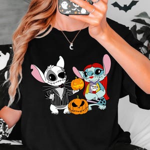 May include: Black t-shirt with a Halloween graphic. The design features cartoon characters dressed as Jack Skellington and Sally from The Nightmare Before Christmas, holding pumpkins. The shirt is worn by a person.