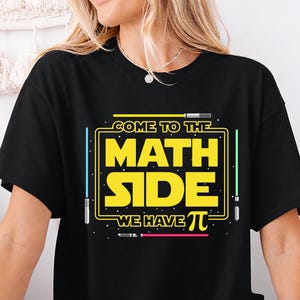 Op de afbeelding: Zwart T-shirt met een geel en witte opdruk met de tekst "COME TO THE MATH SIDE WE HAVE π". Het ontwerp is omlijnd met gele lijnen en bevat lichtzwaard-achtige elementen in blauw en groen.