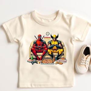 May include: A white t-shirt with a graphic of Deadpool and Wolverine playing video games on a couch. The graphic includes pizza, video game controllers, and the text "Deadpool vs Wolverine".