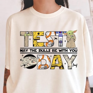 May include: Cream-colored t-shirt featuring the text "TEST DAY" with Star Wars characters integrated into the letters. The phrase "MAY THE SKILLS BE WITH YOU" is below the main text, with a Millennium Falcon graphic and a TIE fighter.