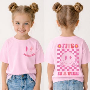 Op de afbeelding: Roze t-shirt met een smiley en bliksem ontwerp. De voorkant van het shirt heeft de naam "Jennifer" afgedrukt. De achterkant van het shirt heeft de tekst "FIVE IS A VIBE" met een smiley en vredestekens.