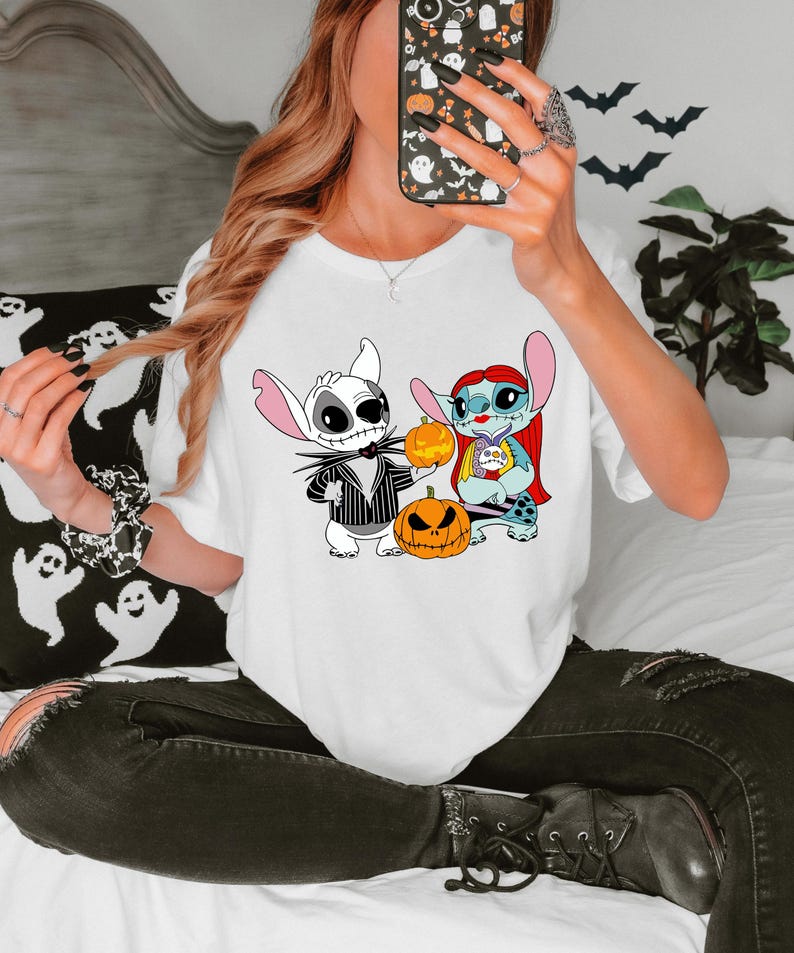 May include: White t-shirt with a Halloween graphic. The design shows two cartoon characters, one dressed as Jack Skellington and the other as Sally, holding pumpkins. Bats are visible in the background.