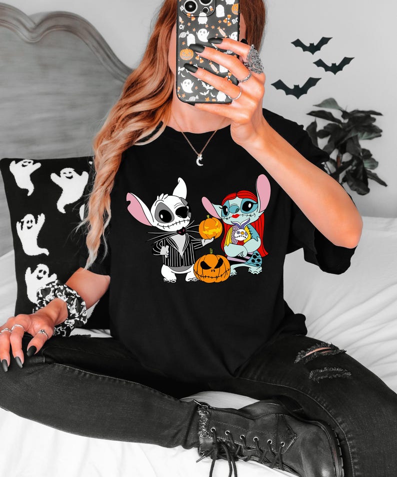 May include: Black t-shirt with a Halloween graphic. The design features cartoon characters dressed as Jack Skellington and Sally from The Nightmare Before Christmas, holding pumpkins. The shirt is worn by a person.