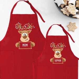 May include: Red Christmas aprons featuring a reindeer design with string lights on the antlers. One apron says "MOM" and the other says "SOPHIA". Both aprons have a front pocket and are displayed with Christmas cookies.