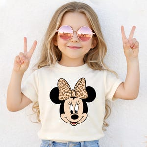 Könnte beinhalten: Ein cremefarbenes T-Shirt mit einem Minnie Mouse-Motiv mit Leopardenmuster-Schleife. Das Bild zeigt eine junge Person mit runden rosa Sonnenbrillen, die mit ihren Fingern ein Friedenszeichen macht.