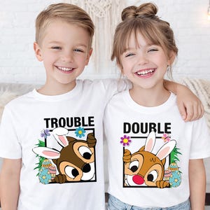 May include: Two children wearing white t-shirts with matching graphics. The shirts feature a cartoon chipmunk wearing bunny ears, with the words "TROUBLE" and "DOUBLE" in black block letters. Easter-themed design with colorful eggs and flowers.