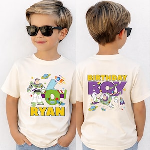 May include: Cream-colored t-shirt featuring a Buzz Lightyear graphic with the number 6 and the name "Ryan" in yellow. The back of the shirt says "Birthday Boy" with a Buzz Lightyear graphic. The shirt is worn by a child.