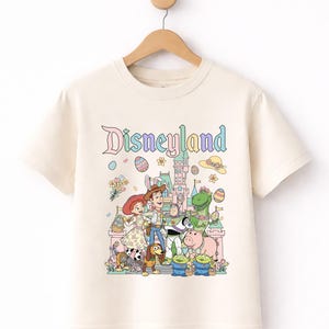 Può includere: T-shirt color crema con grafica colorata di Disneyland. Il design include personaggi dei cartoni animati, un castello e la scritta "Disneyland" in colori pastello. La maglietta è appesa a un appendiabiti di legno.